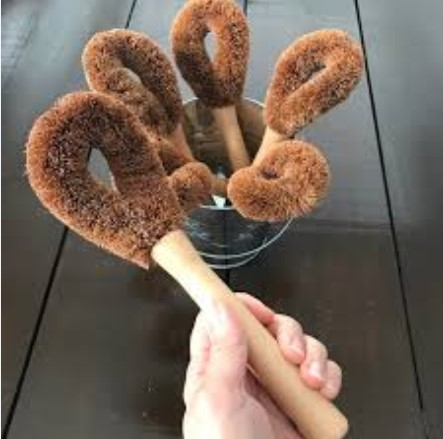 A scouring brush is held in a hand while a collection of scouring brushes suitable for dishes or cleaning toilets sit in an aluminum pail in the background scouring brushes