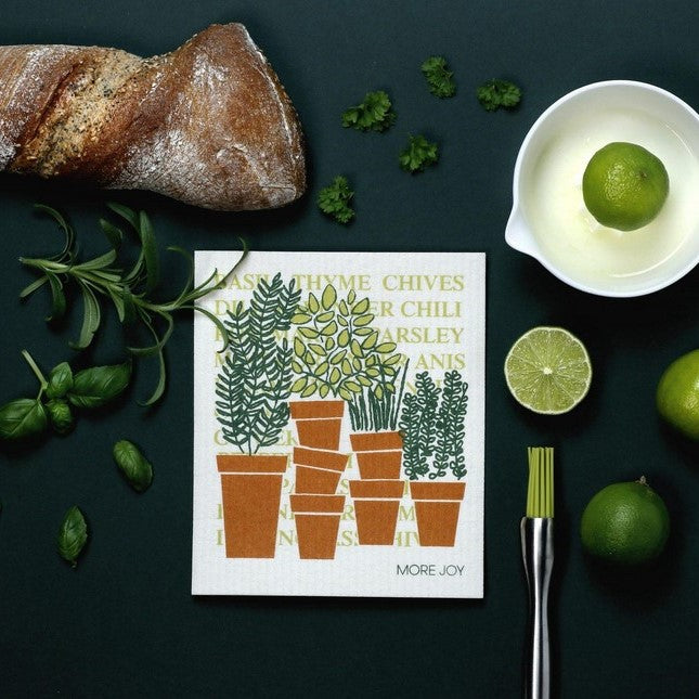A more joy swedish dish cloth with a herb in pots pattern sits on a counter with a loaf of bread, a variety of herbs and some limes