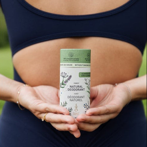 The back of a woman in a navy blue sports bra and matching leggings holds a natural deodorant stick behind her back in both hands. The product packaging is white with a light green top section and botanical illustrations.