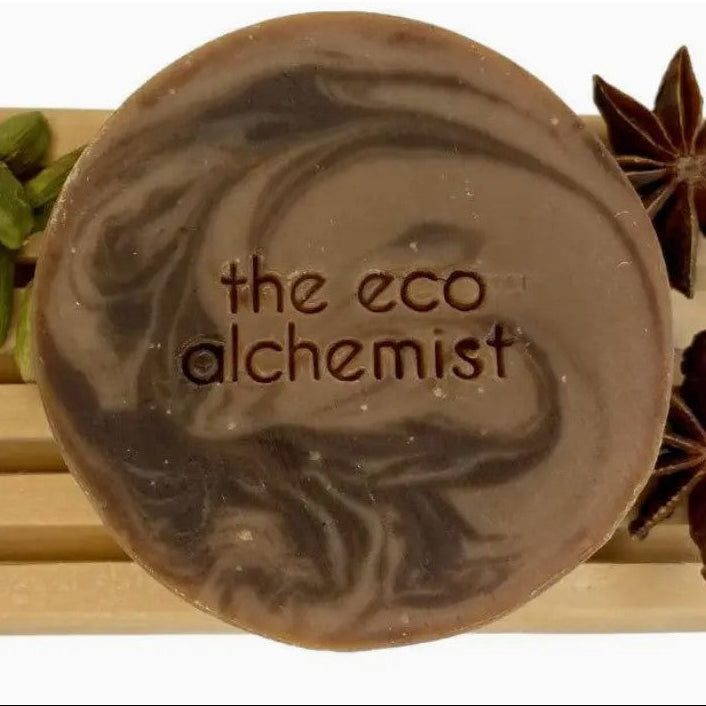 A round artisan soap bar with a dark brown and light brown marbled swirl pattern, featuring the text 'the eco alchemist' embossed in the center. The soap is displayed on a light wooden slatted surface, flanked by decorative star anise and green cardamom pods.
