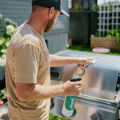 Person cleaning a stainless steel barbecue with myni Degreaser Spray in a backyard, showcasing an effective plant-based cleaner for removing grease and grime from outdoor grills.