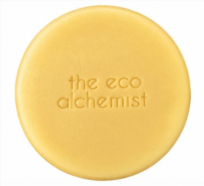 A round, pale yellow bar of soap with the words 'the eco alchemist' embossed in the center, shown against a plain white background.