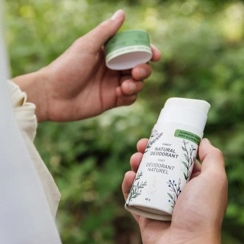A person outdoors holds a white deodorant bottle with green botanical text and graphics, while another hand lifts the green cap off the bottle. The background is a soft-focus green natural setting.
